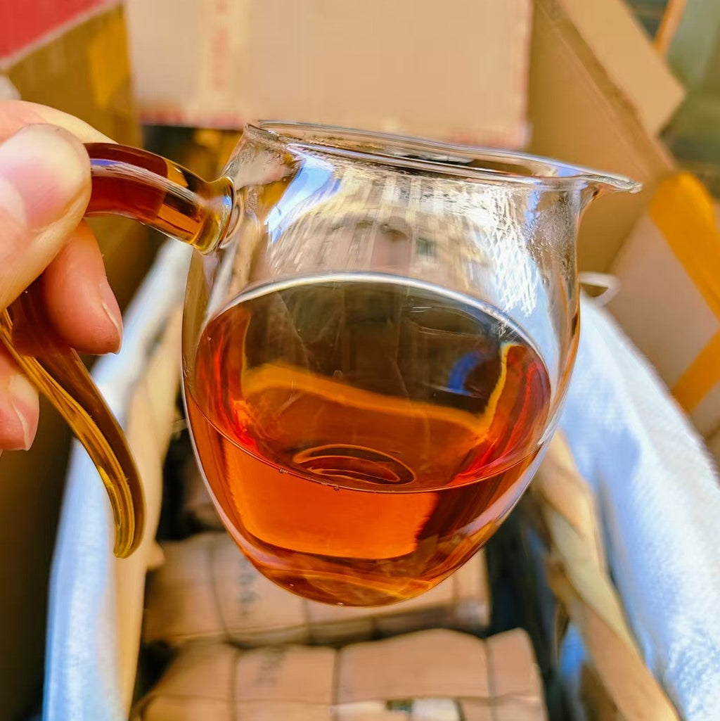 Glass teacup with a handle filled with tea, held by a hand against a cardboard box background.