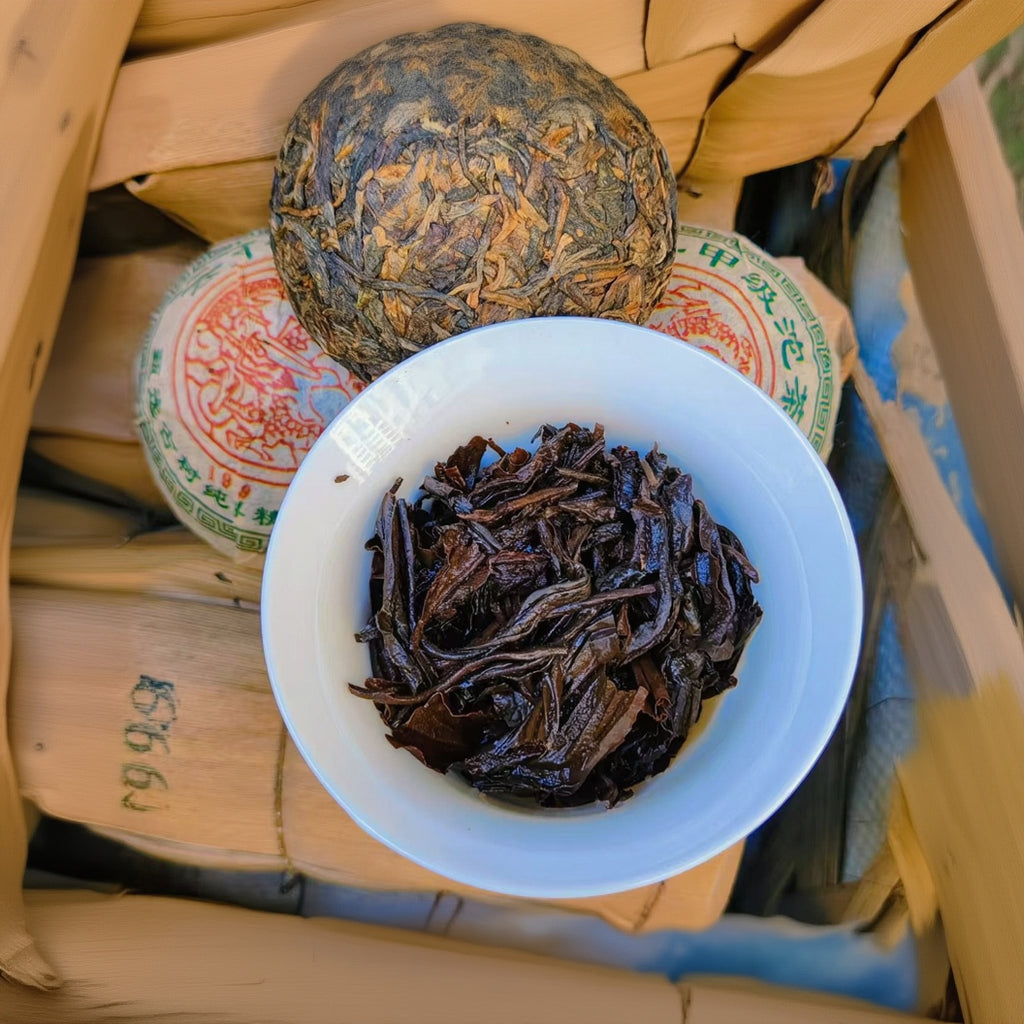 White bowl filled with dark tea leaves on wooden boxes with tea packages.