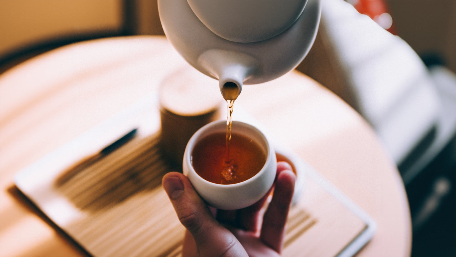 Tea being poured from a white teapot into a white cup on a wooden table.